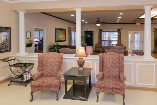 Two patterned armchairs flank a small table with a lamp in a furnished common living area framed by white columns and additional seating in the background.