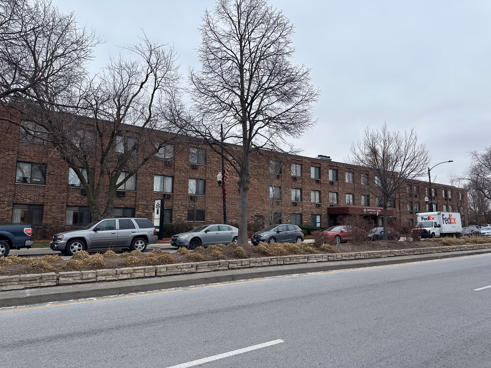 Exterior view of a three-story brick building with multiple windows, several parked cars, and two FedEx trucks along the street. Leafless trees and a stone-lined median are visible in front of the building under an overcast sky.