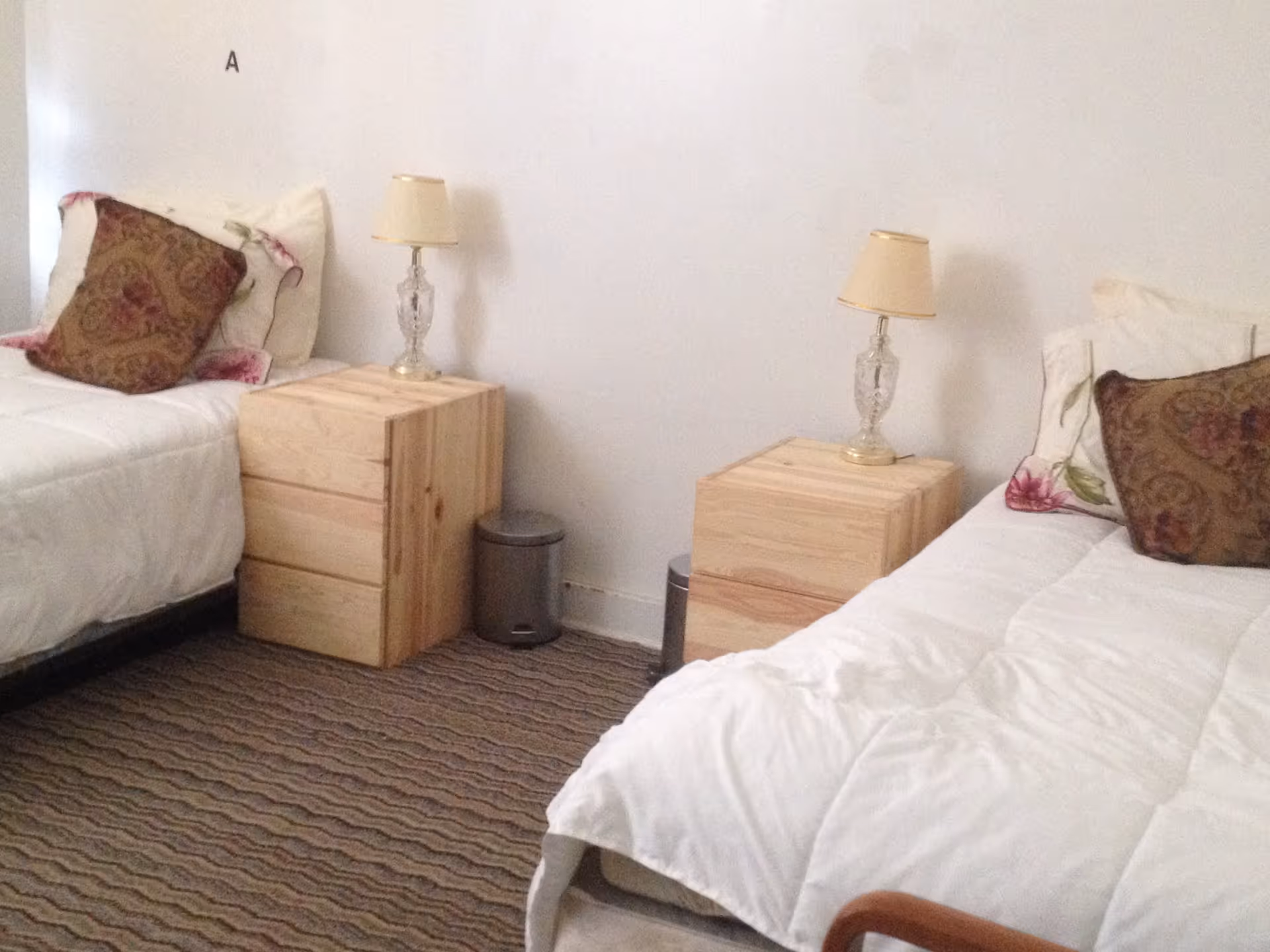 A bedroom with two single beds separated by two wooden nightstands. Each nightstand has a small lamp with a beige lampshade. Both beds have white bedding and decorative pillows with a floral pattern. The floor is carpeted with a wavy pattern, and there is a small trash can between the nightstands against a plain white wall.