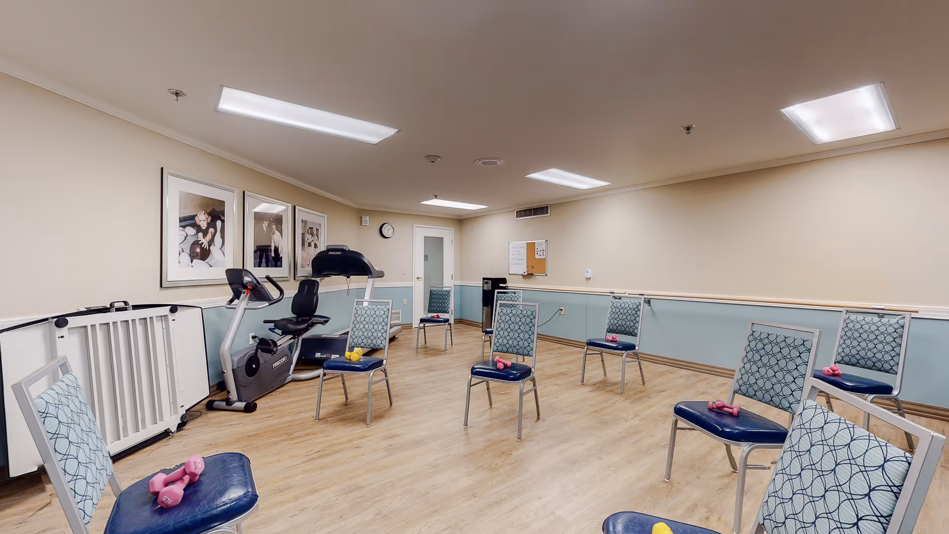A senior activity/exercise room with spaced chairs holding small dumbbells and a stationary bike along the wall.