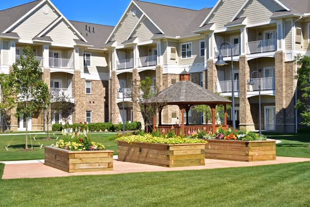 Outdoor garden area with raised wooden flower beds and a wooden gazebo in front of a multi-story senior living building with balconies under a clear blue sky.