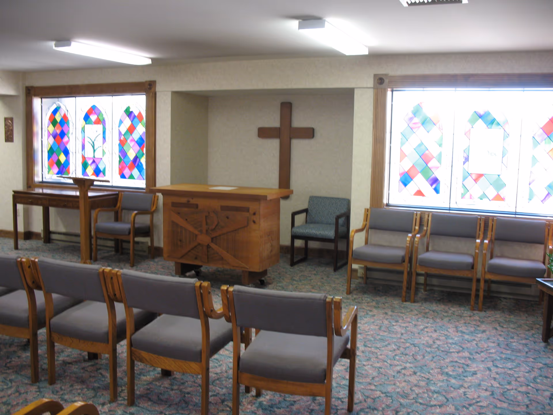 Interior room with rows of wooden chairs with gray cushions facing a wooden podium and a wooden cross mounted on the wall. The room has stained glass windows with colorful diamond patterns on both sides.