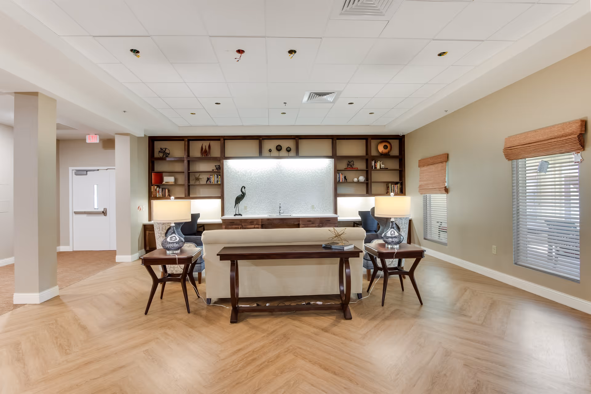 A bright and spacious living room area in an assisted living facility featuring a beige sofa with a wooden console table behind it, two side tables with lamps, two blue armchairs, and a large built-in wooden shelving unit with decorative items and books. The room has light wood flooring, beige walls, and two windows with brown blinds.