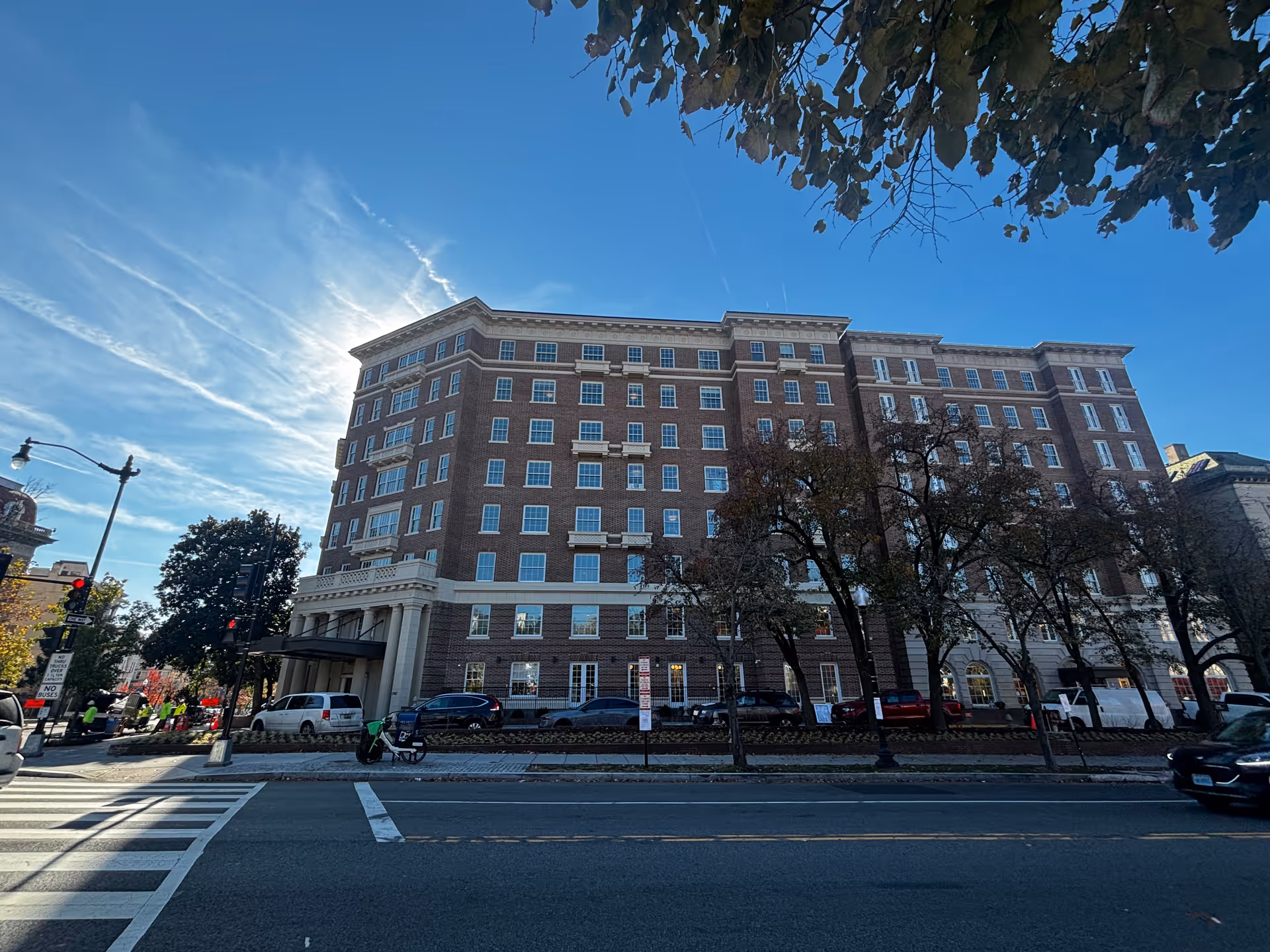 Front exterior of a multi-story brick residential building with trees, parked cars, and a crosswalk in front.