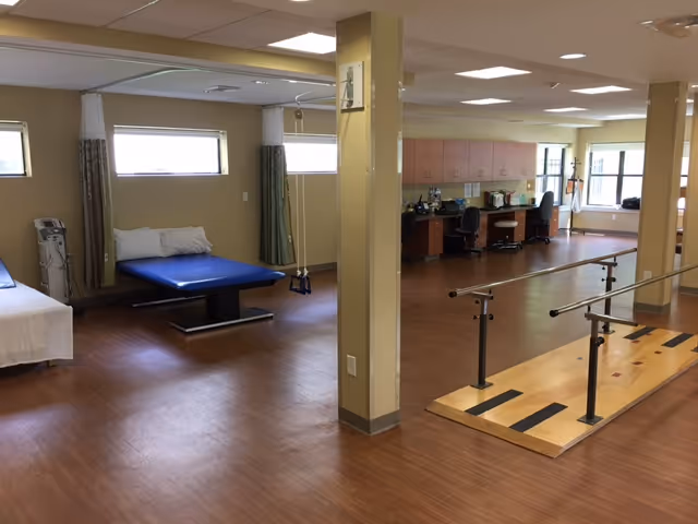 Rehabilitation therapy room with parallel walking bars, treatment tables, and workstations on wood-look flooring.