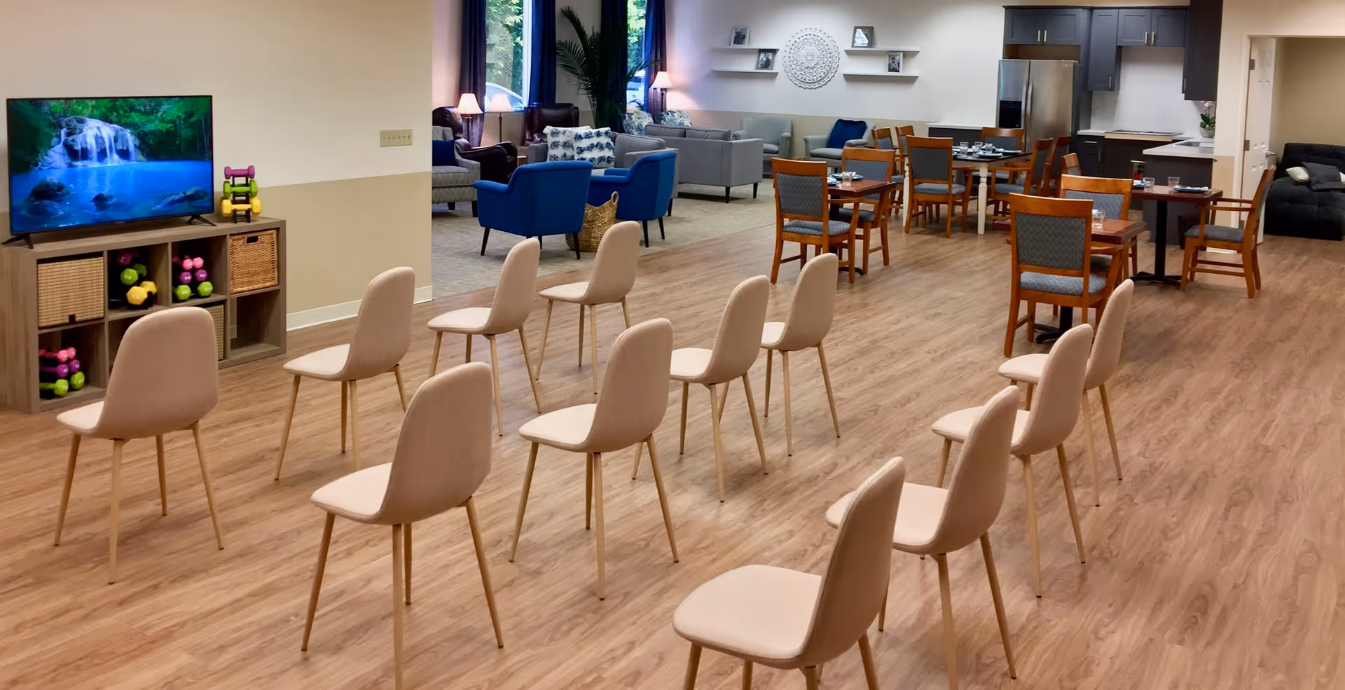 A spacious room with multiple beige chairs arranged in rows facing a television displaying a waterfall scene. Behind the chairs, there is a seating area with blue and gray armchairs and sofas, a dining area with wooden tables and chairs, and a kitchen area with dark cabinets and a stainless steel refrigerator. The room has wood flooring and large windows with blue curtains.