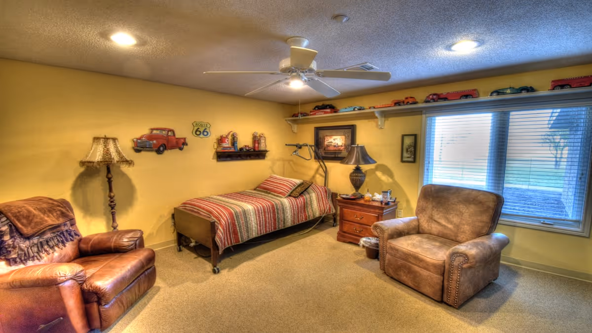 A cozy room with yellow walls featuring a single bed with a striped blanket, two brown leather armchairs, a wooden nightstand with a lamp, and a window with blinds letting in natural light. The walls are decorated with vintage car-themed items and a Route 66 sign. A ceiling fan with a light is mounted on the ceiling.