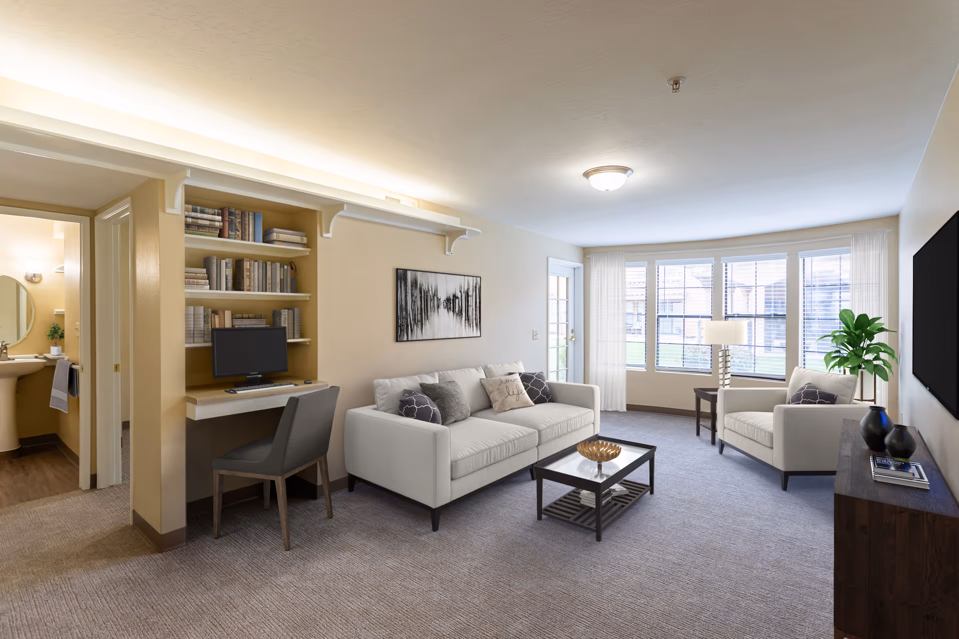 Bright furnished living room with a sofa, armchair, coffee table, desk with computer and built-in shelves, and large windows.