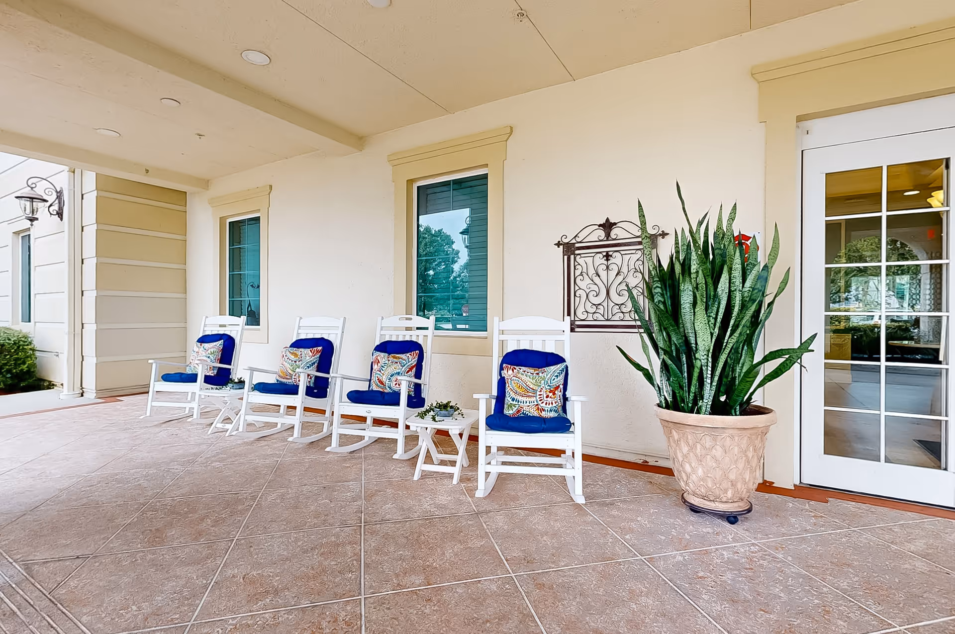 Covered outdoor patio area with four white rocking chairs, each with a blue cushion and a colorful decorative pillow. There is a small white side table with a plant on it between the chairs. A large potted plant is placed near a glass door leading inside the building. The walls are light-colored with two windows and decorative metal wall art.