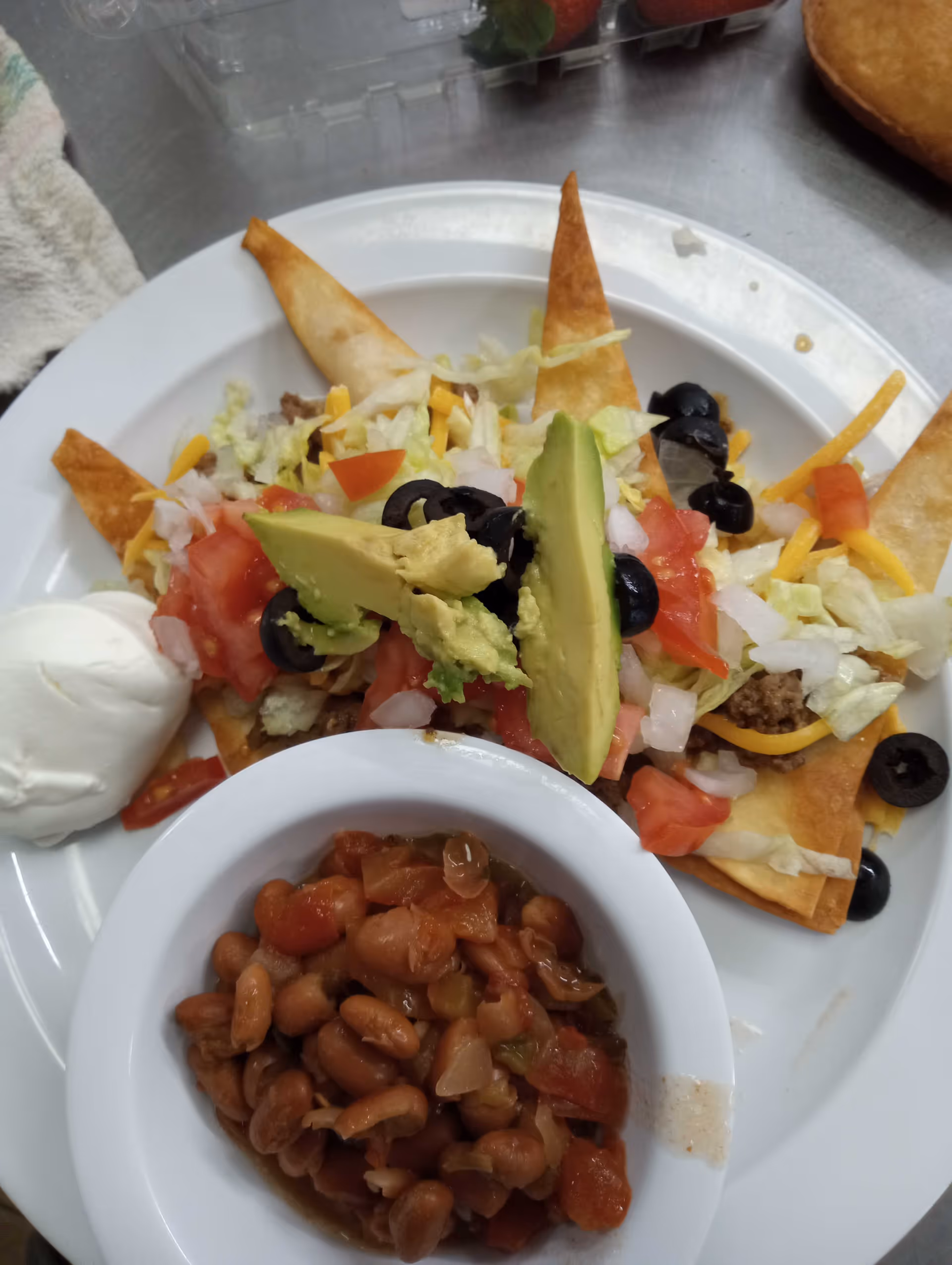 A plate of nachos topped with ground beef, shredded lettuce, diced tomatoes, sliced black olives, shredded cheddar cheese, and avocado slices, served with a dollop of sour cream on the side. A small bowl of cooked pinto beans with diced tomatoes and peppers accompanies the nachos on the plate.