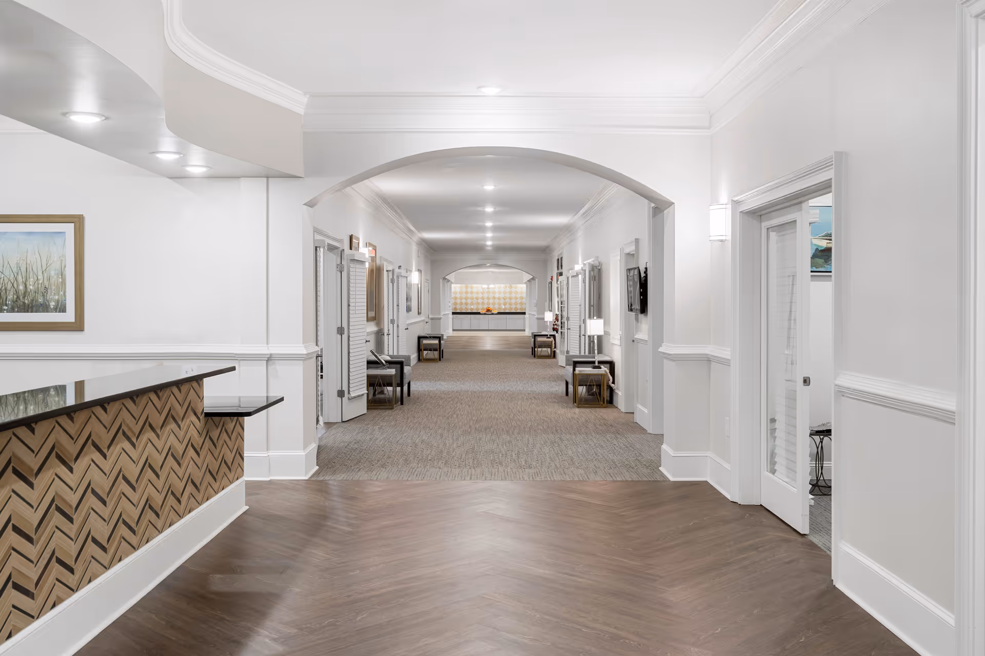 A spacious, well-lit hallway in a senior living facility with a reception desk on the left featuring a chevron wood pattern and a black countertop. The hallway has white walls with crown molding, multiple doors on both sides, and several small tables with lamps and chairs along the walls. The floor transitions from wood near the reception area to carpet further down the hallway.