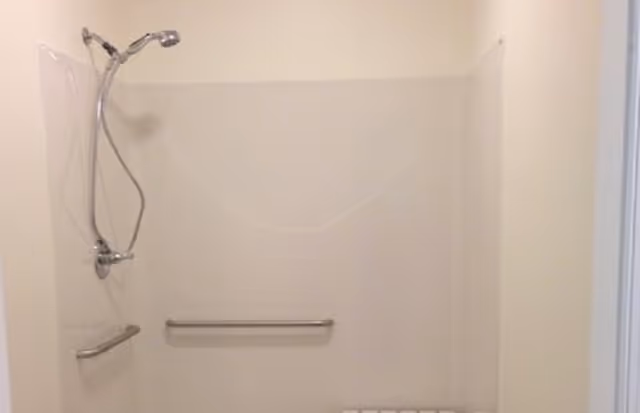 A simple shower area with a handheld showerhead attached to a wall mount, two horizontal grab bars on the walls, and a white wall surround.