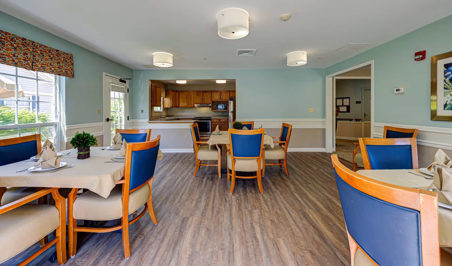 A dining room in a senior living facility with several tables covered with beige tablecloths, each set with plates, napkins, and silverware. The chairs have wooden frames with blue cushioned backs and beige seats. The room has light blue walls with white trim, wood-look flooring, and three ceiling light fixtures. A kitchen area with wooden cabinets and appliances is visible through an open wall space. A large window on the left lets in natural light, and a door is next to it.