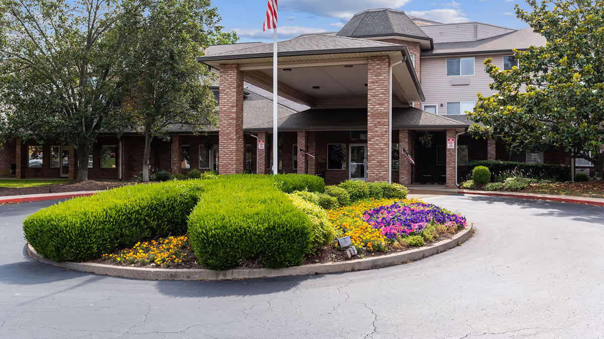 Front entrance of Holiday Uffelman Estates featuring a covered drop-off area with brick pillars, a circular driveway, well-maintained green shrubs, colorful flower beds, and an American flag on a flagpole. Trees and a partly cloudy sky are visible in the background.