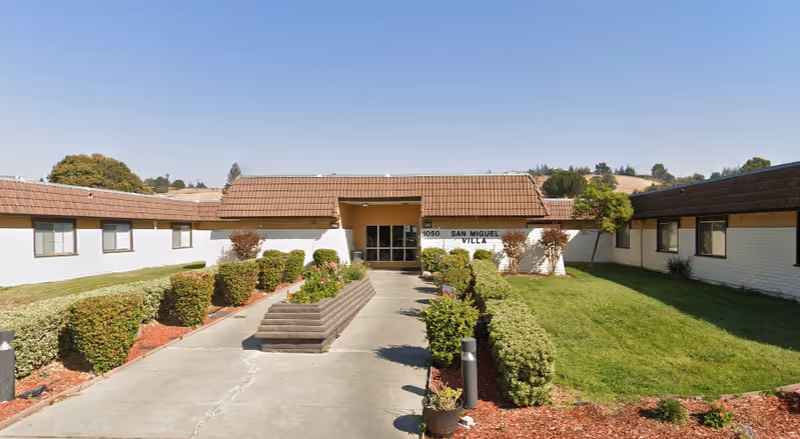 Exterior view of a single-story building with a brown tiled roof and white walls, surrounded by neatly trimmed bushes and green grass under a clear blue sky. A concrete pathway leads to the entrance with glass doors. The building has a sign that reads '1050 San Miguel Villa'.