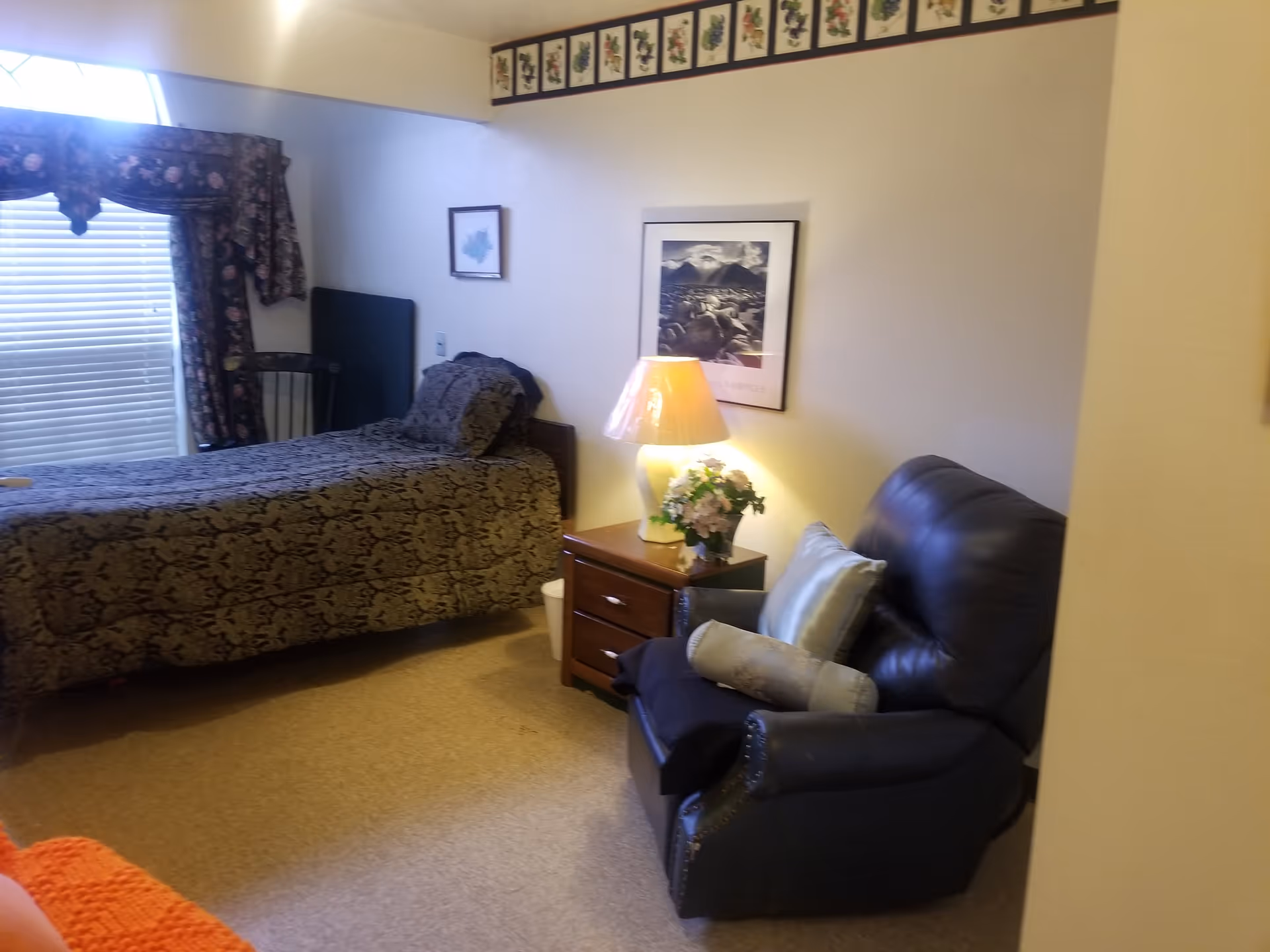 Small furnished bedroom with a single bed, bedside table and lamp, framed art, and a recliner chair.