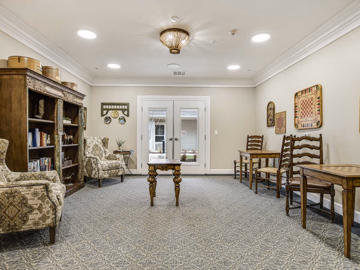 A cozy common area with patterned carpet, two upholstered armchairs, a wooden bookshelf filled with books and decorative items, a small wooden table in the center, and two wooden tables with chairs along the right wall. The room has beige walls, a ceiling light fixture, recessed lighting, and double glass doors leading outside.