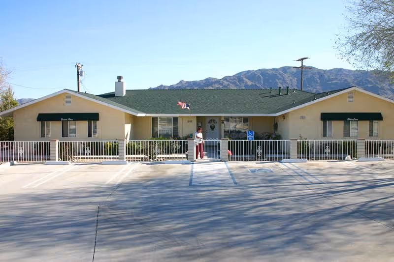 Single-story building with a green roof and beige walls, featuring two green awnings on either side labeled Desert Rose. A person stands near the entrance gate in front of the building, with a mountain range visible in the background under a clear blue sky. The foreground shows an empty parking lot with a handicap parking sign near the entrance.