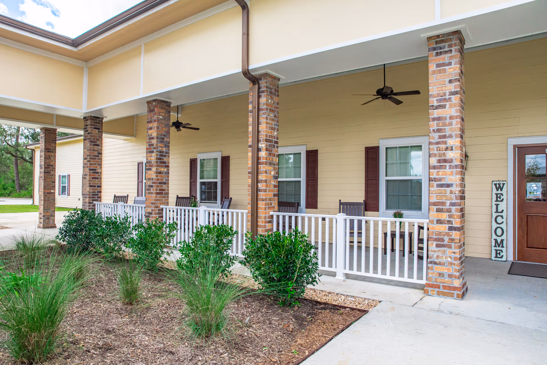 Covered porch area of a senior living facility with brick columns, white railing, several rocking chairs, ceiling fans, and a welcome sign near a wooden door. There are shrubs and plants in the garden bed in front of the porch.