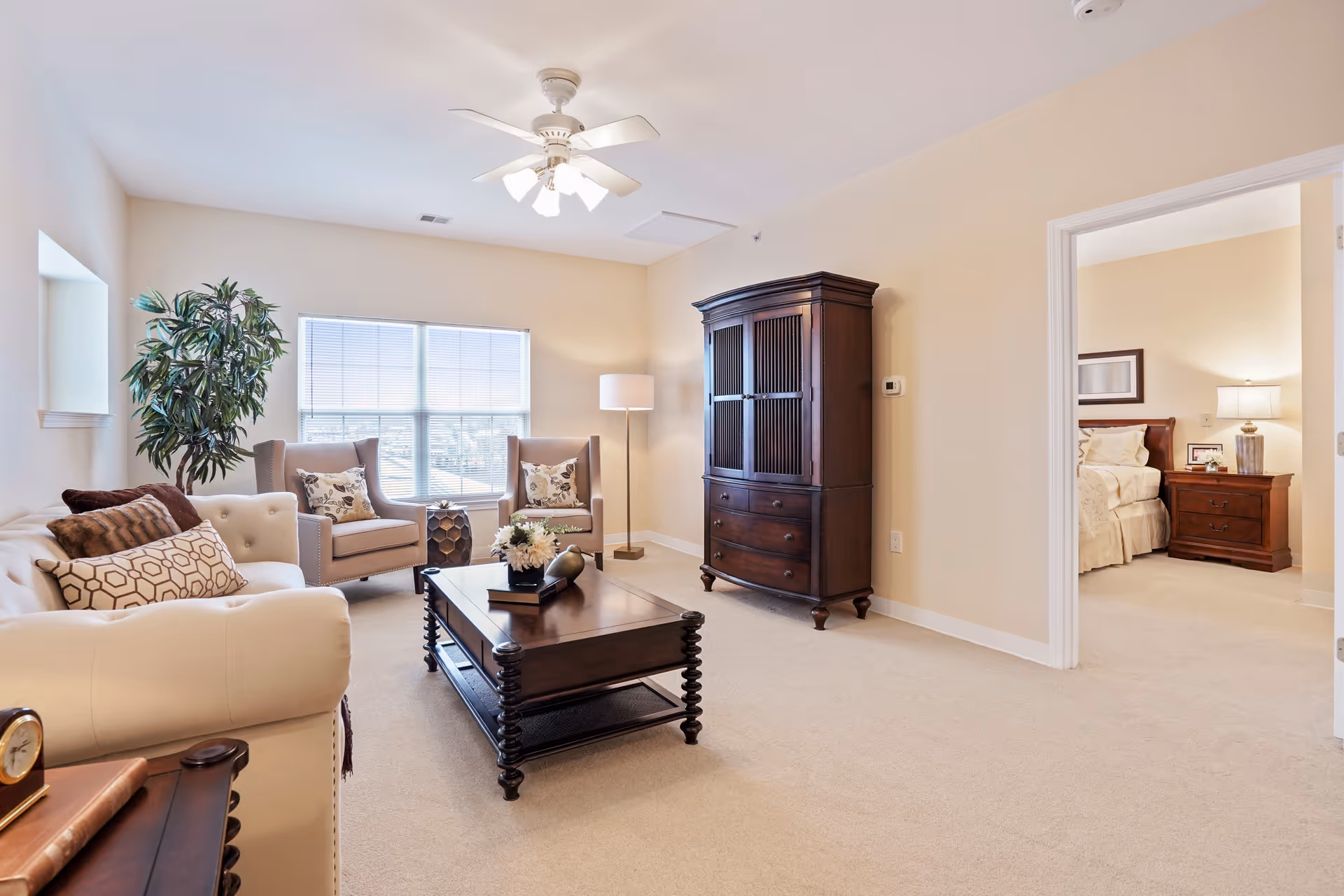 A bright and cozy living room with a white sofa, two armchairs with patterned pillows, a dark wooden coffee table with decorative items, a tall dark wooden cabinet, a floor lamp, and a large window letting in natural light. An open doorway reveals a bedroom with a bed, nightstand, and lamp.