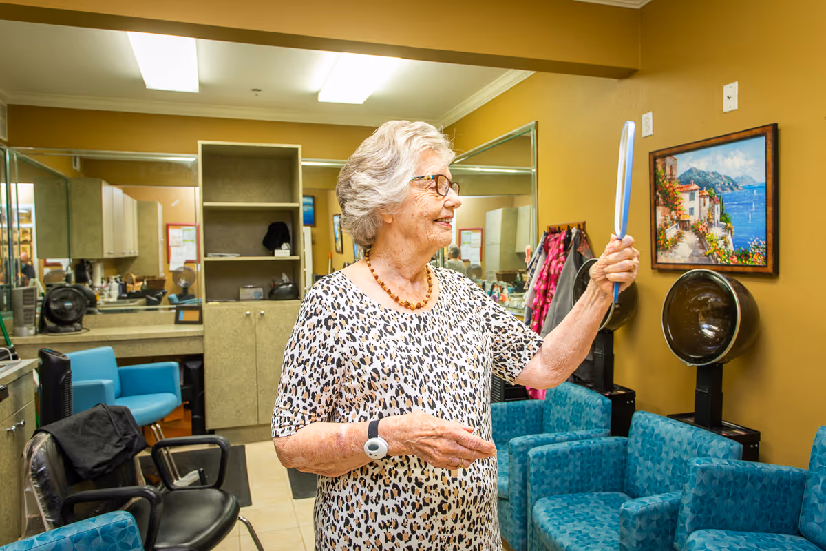 An elderly woman with gray hair and glasses stands in a salon area holding a hand mirror and smiling. The room has mustard yellow walls, blue salon chairs, hair drying stations, and a painting of a coastal scene on the wall. There are mirrors and cabinets in the background.