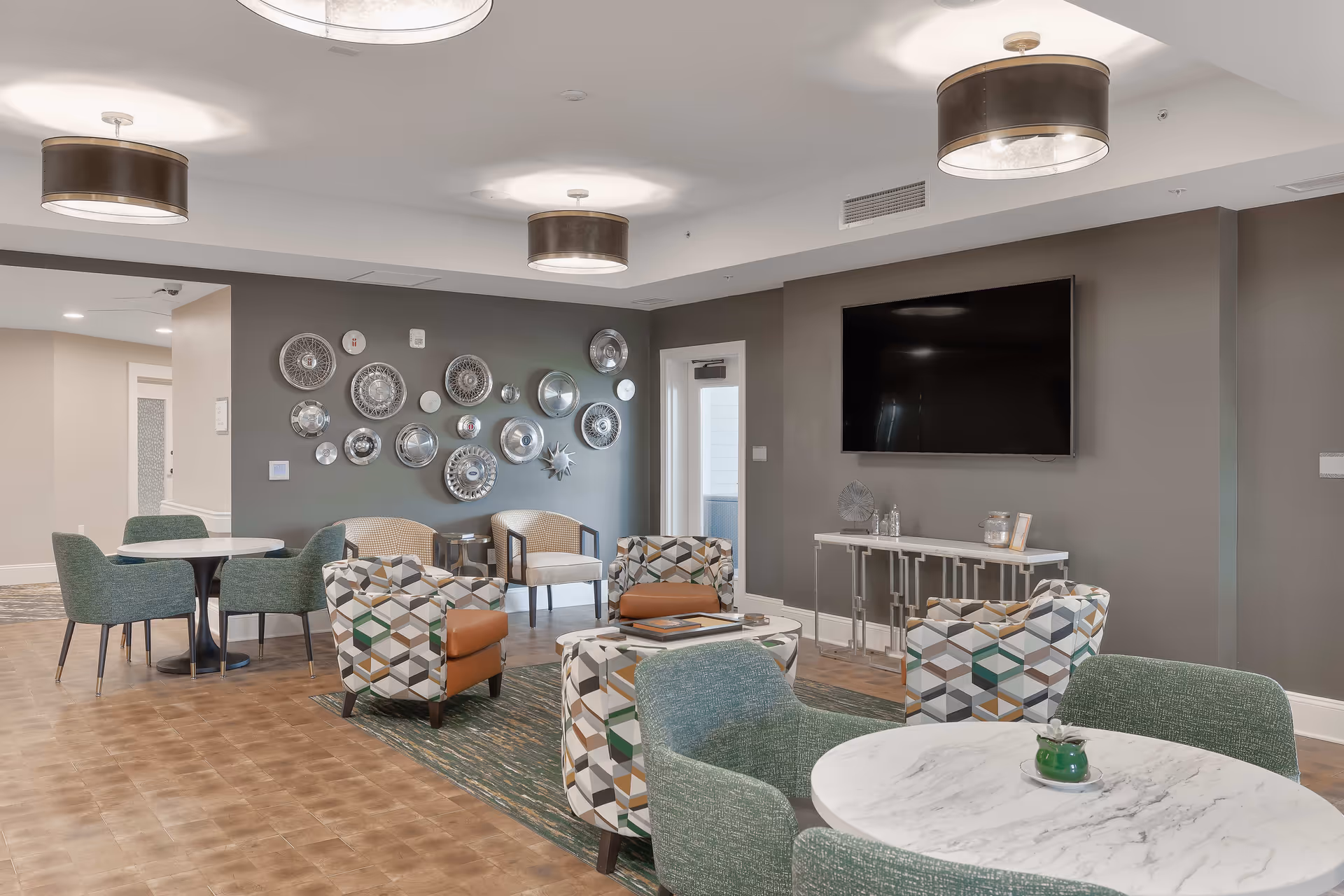 A modern living room area in a senior living facility with patterned armchairs, green upholstered chairs around white marble tables, a large flat-screen TV mounted on a gray wall, and decorative hubcaps arranged on another wall. The room has wooden flooring, a green rug, and three ceiling light fixtures.