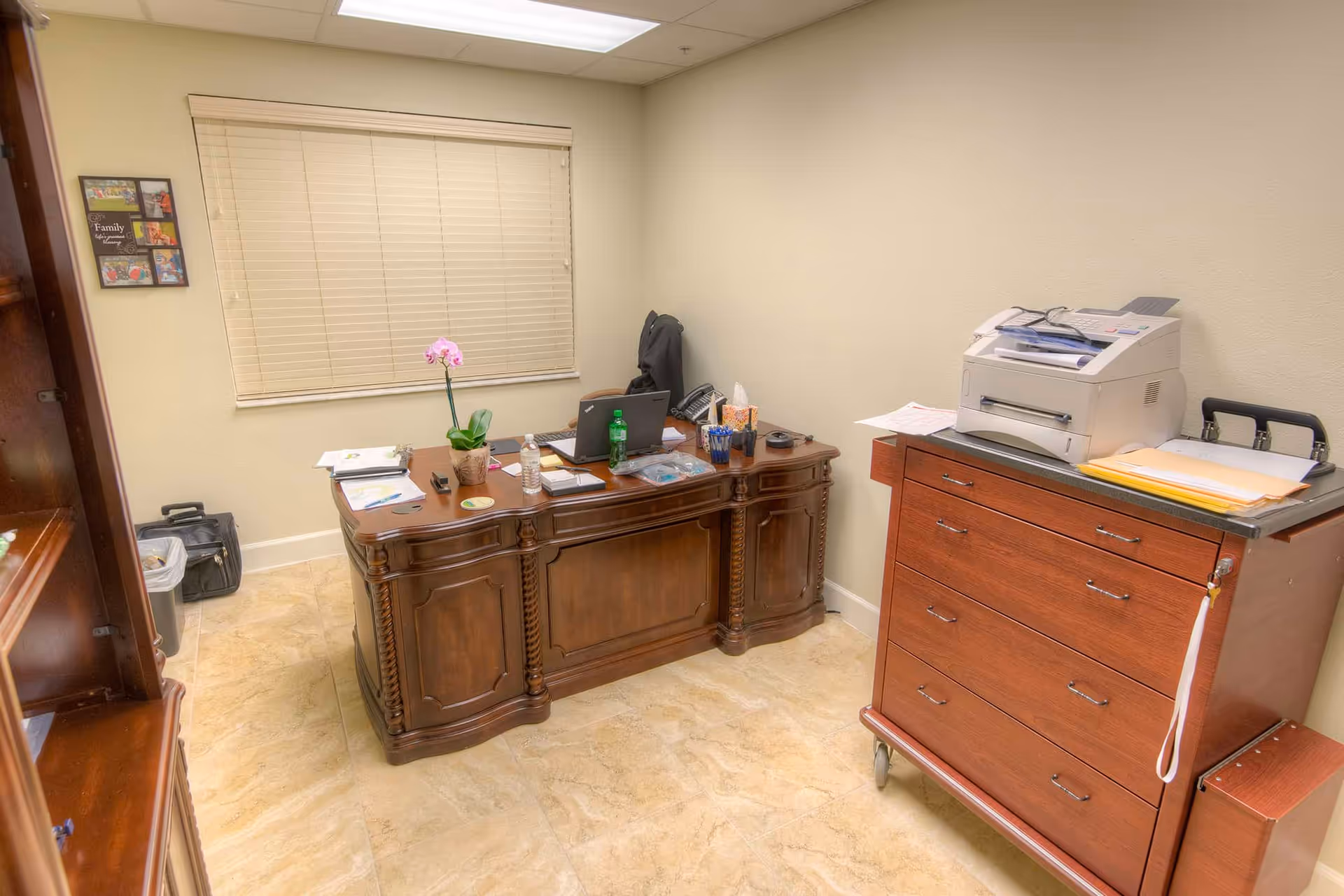 A small office room with a wooden desk holding a laptop, water bottle, soda bottle, tissue box, and various papers. There is a wooden cabinet with drawers on wheels to the right, topped with a printer and more papers. A window with closed blinds is on the back wall, and a framed collage hangs on the left wall. The floor is tiled and the walls are painted beige.