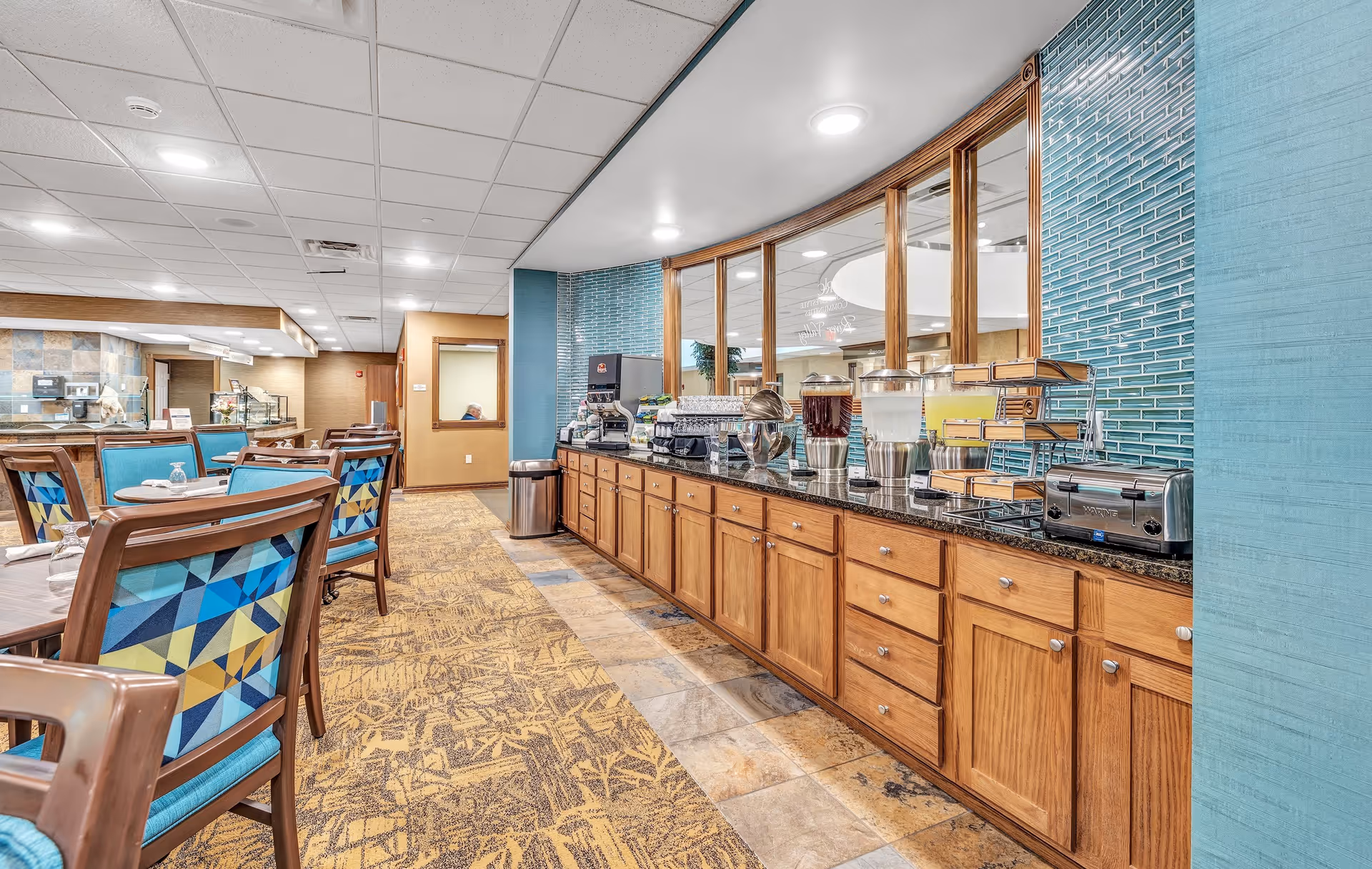 Interior view of a dining area at River Valley Retirement Community featuring wooden tables and chairs with colorful geometric patterned cushions. On the right side, there is a long counter with wooden cabinets below and a black granite countertop holding beverage dispensers, a toaster, and a coffee machine. The wall behind the counter has blue tile accents and large windows looking into another room.
