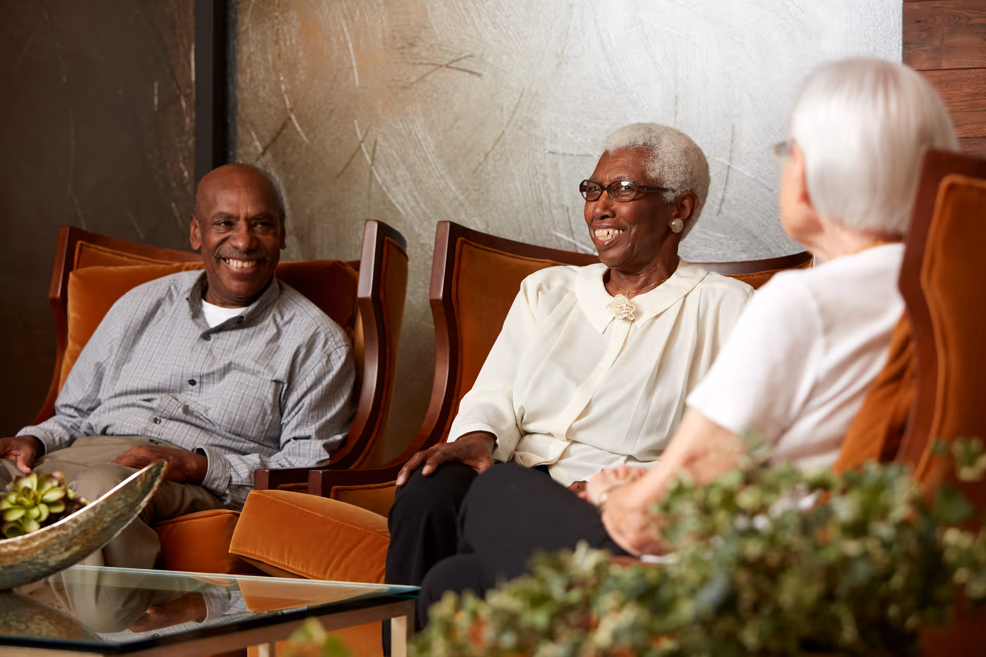 Three elderly individuals sitting and smiling in a cozy living room area with orange cushioned chairs and a glass coffee table with a decorative bowl and plant in the foreground.