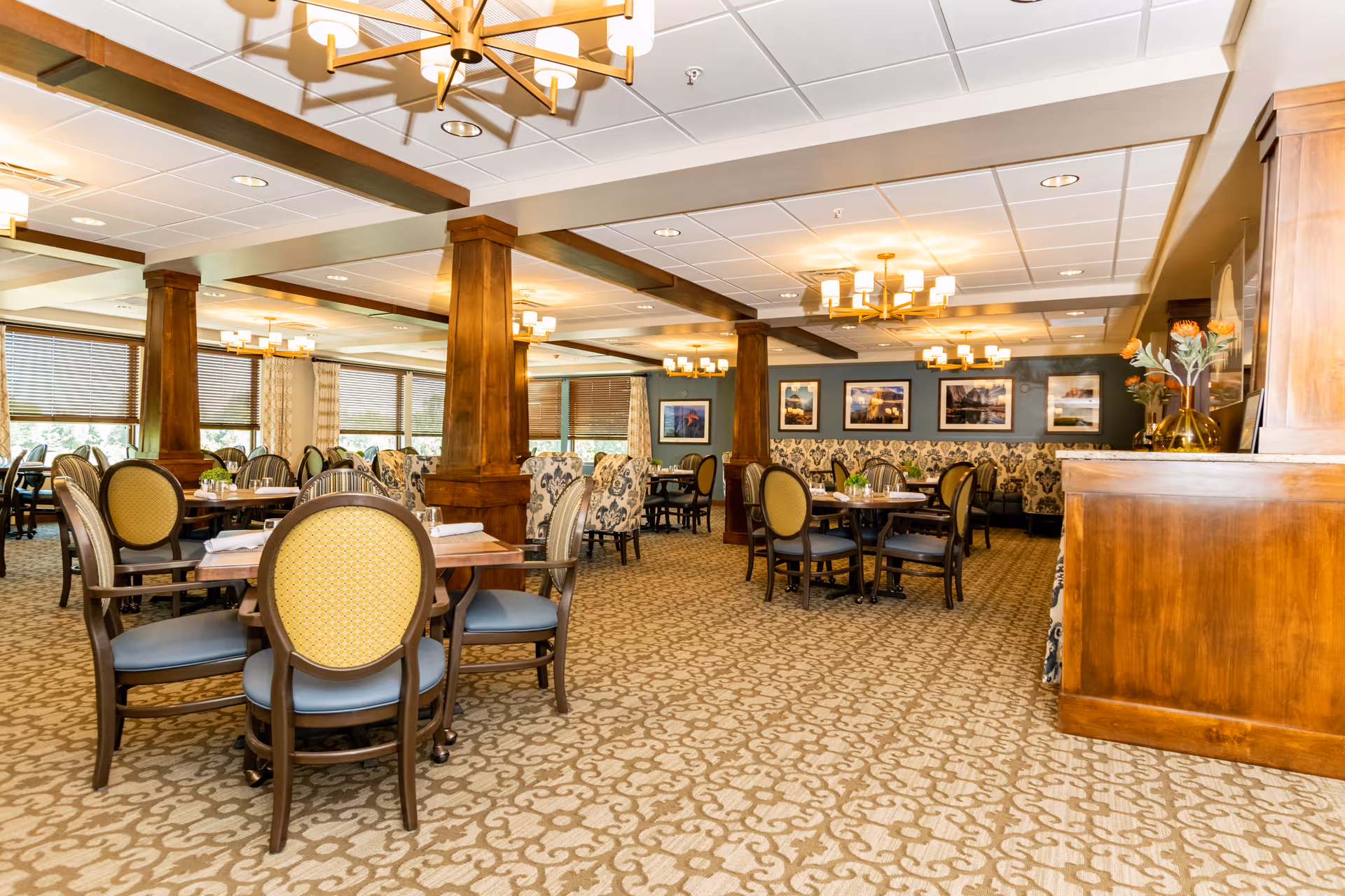 A spacious dining room with multiple round wooden tables and cushioned chairs arranged neatly. The room features wooden columns, patterned carpet, large windows with blinds, and framed pictures on the walls. Warm lighting fixtures hang from the ceiling, creating a cozy atmosphere.