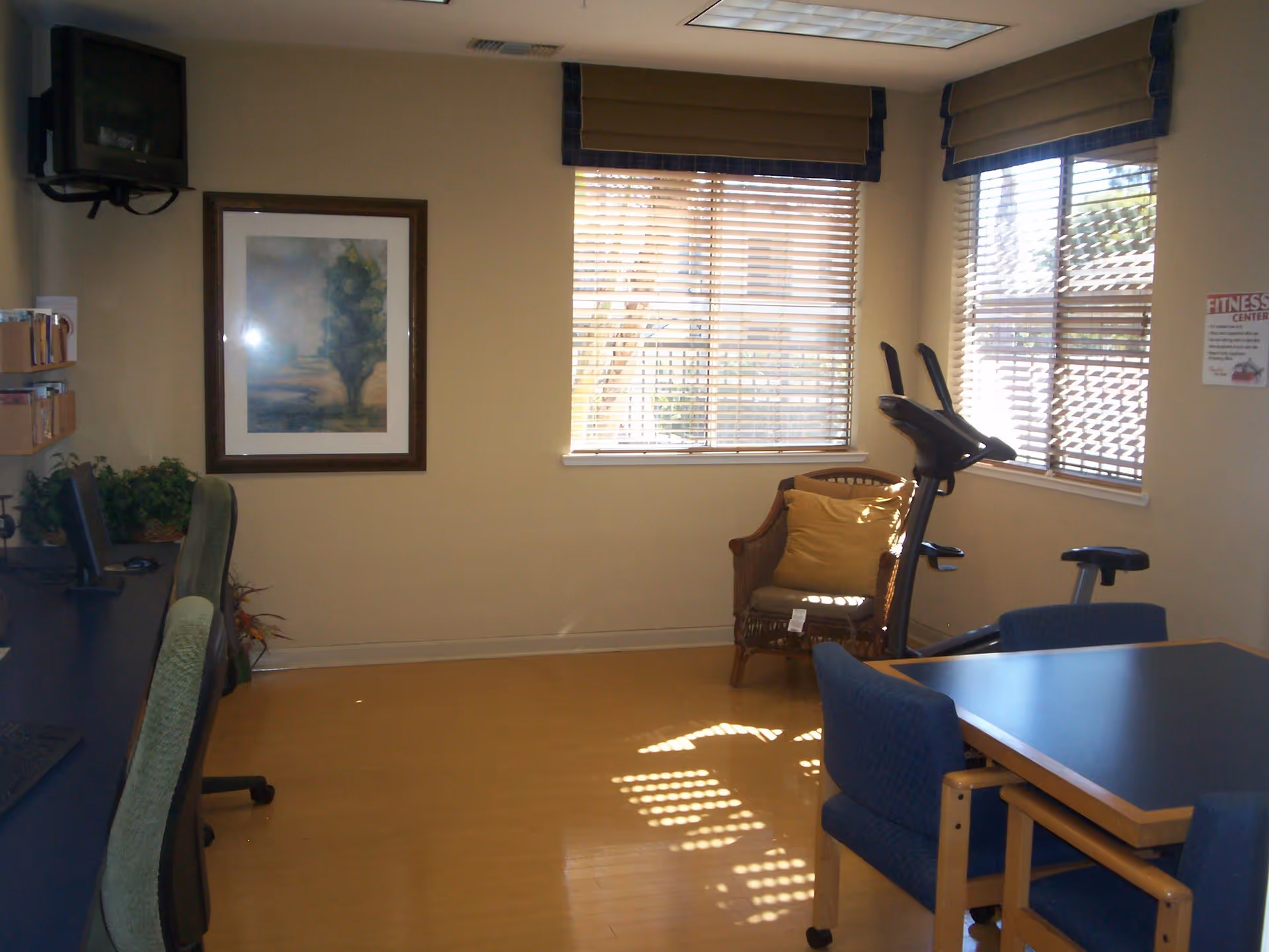 A small room with two windows covered by blinds and valances, a wicker chair with a yellow cushion, an exercise bike, a table with four blue chairs, a wall-mounted TV, a framed painting, and a desk with two green office chairs and a computer.