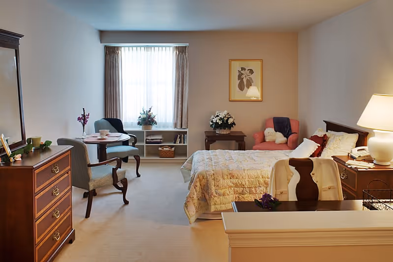 A cozy bedroom in a senior living facility with a bed covered in a floral quilt, a wooden dresser with a mirror, a small round table with two chairs near a window with curtains, a pink armchair with a stuffed animal, a side table with a lamp, and decorative flowers and framed artwork on the walls.