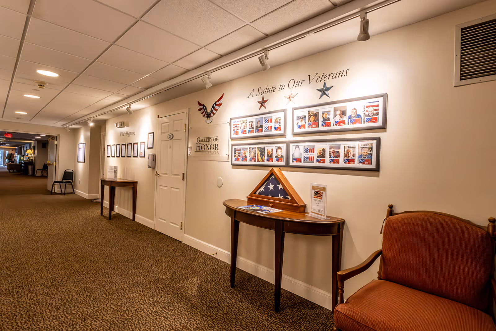 A well-lit hallway in a senior living facility with a carpeted floor and white walls. On the right wall, there is a display titled 'A Salute to Our Veterans' featuring framed photos of veterans, decorative stars, and a folded American flag in a triangular display case on a wooden table. There is also a brown upholstered chair next to the table. Further down the hallway, additional framed pictures and a small table with a chair are visible.