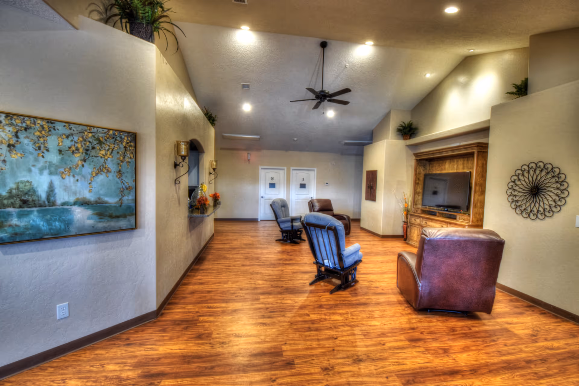 A spacious living room area with wooden flooring, several armchairs arranged facing a wooden entertainment center with a flat-screen TV. The walls are beige with decorative elements including a large painting of a nature scene and a metal wall art piece. The ceiling has recessed lighting and a ceiling fan.