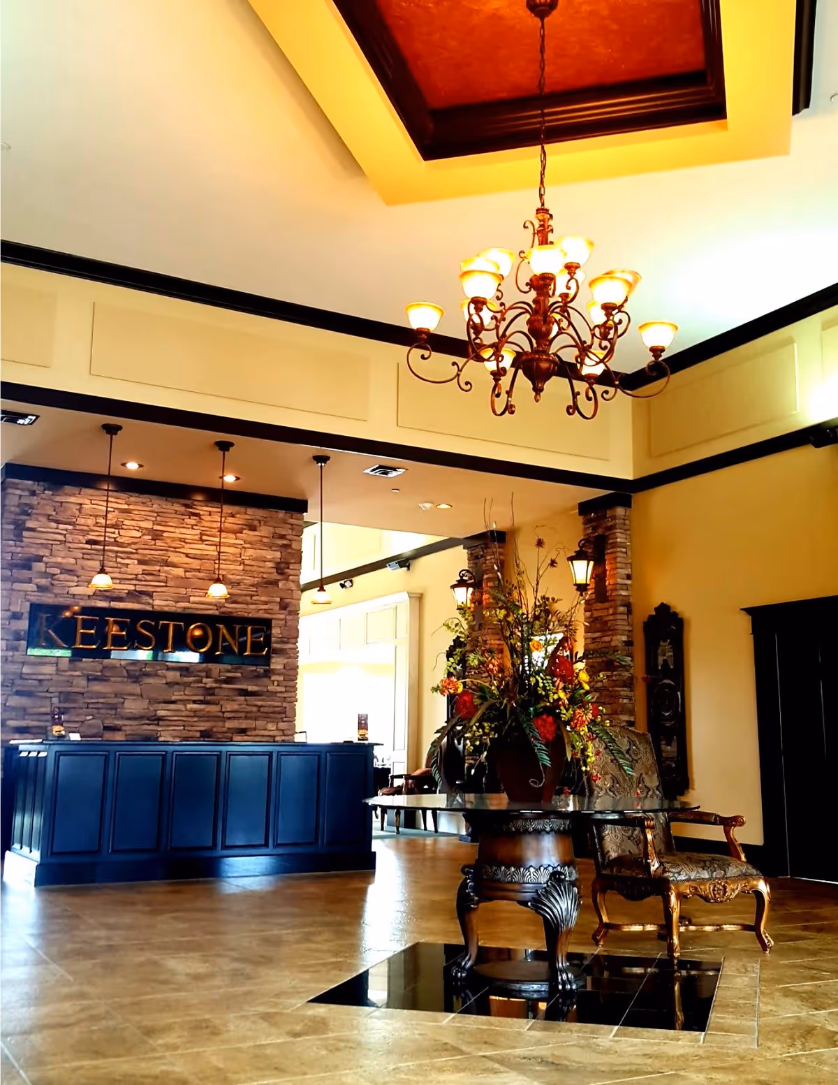 Interior view of a senior living facility lobby with a stone accent wall behind a dark blue reception desk. The wall features a sign that reads 'KEESTONE'. A decorative chandelier hangs from a recessed ceiling with warm lighting. In the foreground, there is an ornate wooden table with a glass top holding a large floral arrangement, and an upholstered armchair with wooden arms and legs is positioned nearby.
