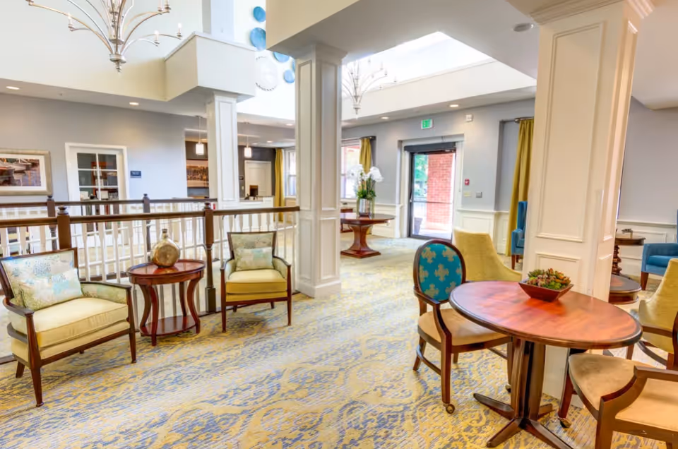 A bright and spacious common area in a senior living facility featuring comfortable armchairs, small wooden tables, decorative vases, and a patterned carpet. Large windows and glass doors allow natural light to fill the room, and elegant chandeliers hang from the ceiling. The space has a welcoming and cozy atmosphere with light-colored walls and tasteful decor.