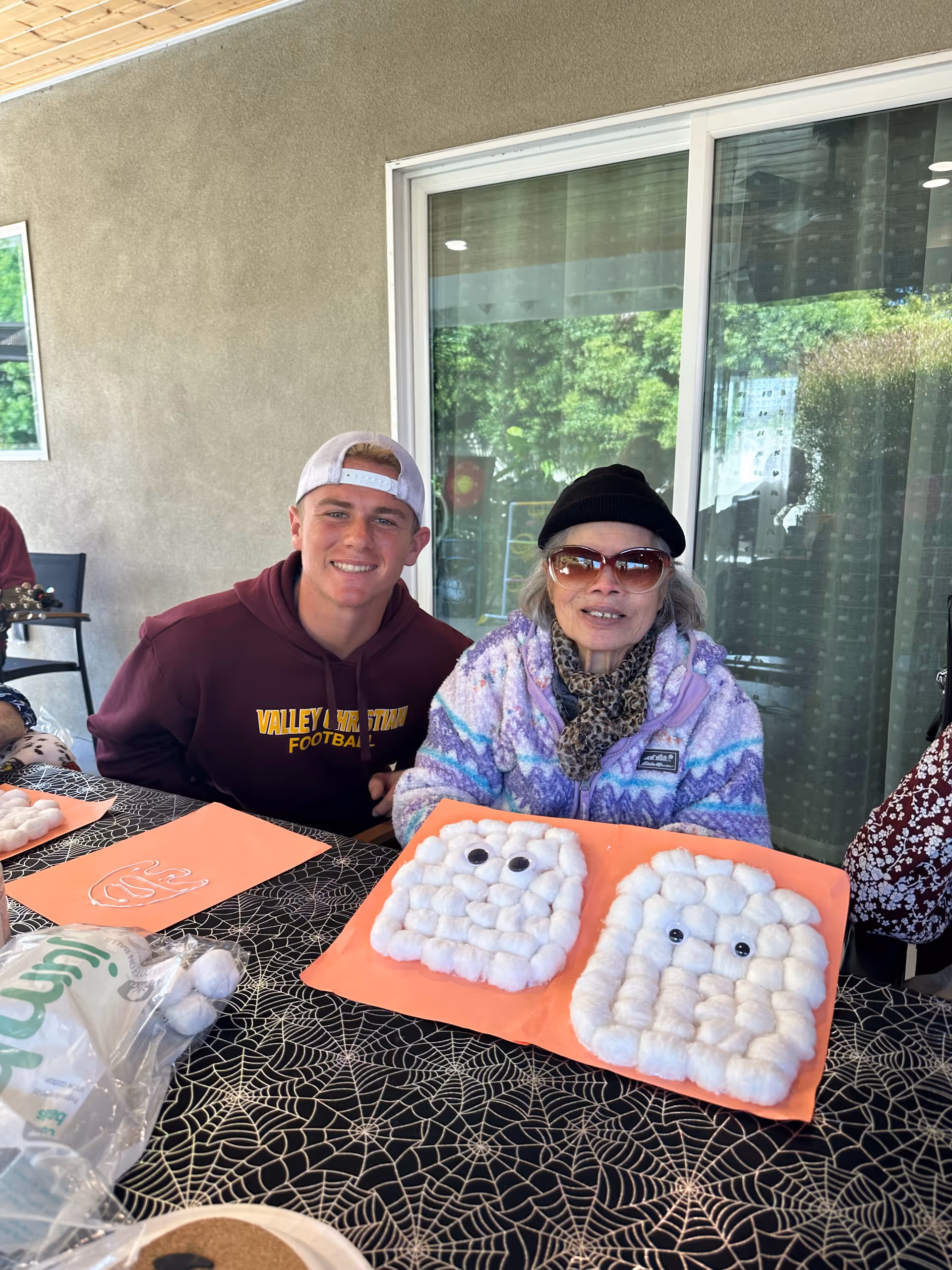 A young man wearing a maroon hoodie and white backwards cap sits next to an elderly woman wearing sunglasses, a black hat, and a colorful jacket. They are seated at a table covered with a black tablecloth featuring a spiderweb pattern. In front of them are two orange sheets with craft projects made of cotton balls arranged to look like ghost faces with googly eyes. The setting appears to be an outdoor covered patio with a sliding glass door and greenery visible outside.
