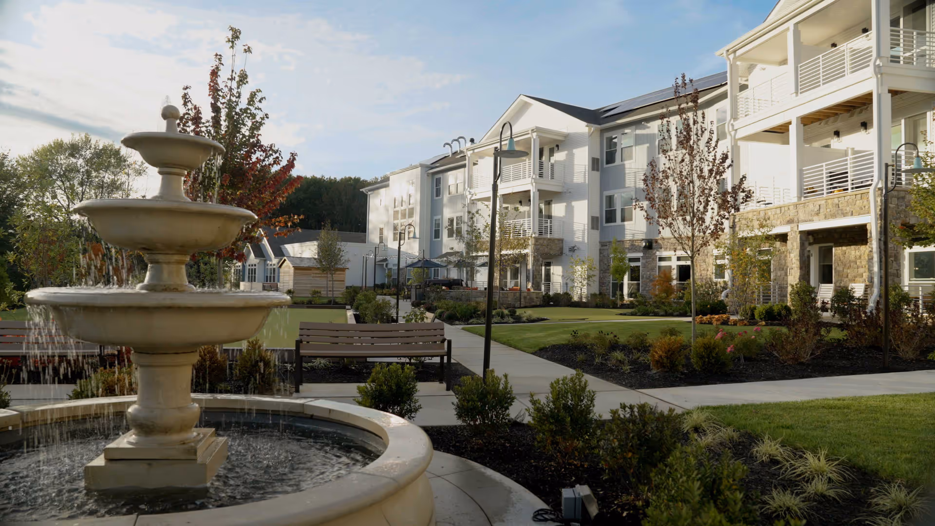 Three-tier stone fountain in a landscaped courtyard with benches and a multi-story senior living building in the background.