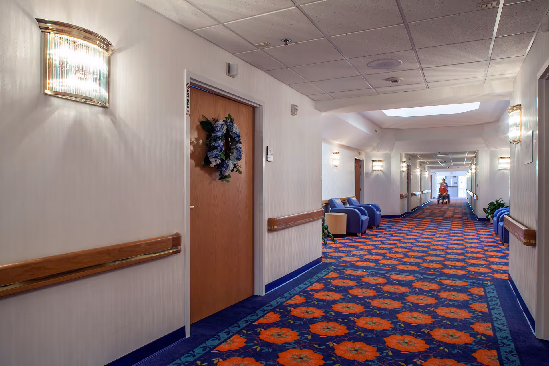A long, well-lit hallway in a senior living facility with patterned carpet featuring orange flowers on a blue background. The walls are light-colored with wooden handrails. There are several wooden doors, one decorated with a blue floral wreath. Blue armchairs and small tables are placed along the hallway. A person in a red shirt is seen in the distance near the end of the hallway.