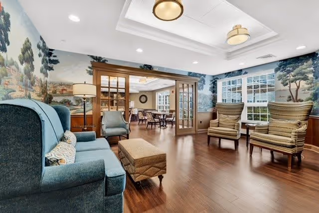 A cozy sitting area in a senior living facility with a blue upholstered sofa, two patterned armchairs, and a cushioned ottoman on a wooden floor. The walls feature a scenic mural with trees and a countryside landscape. Through glass-paneled double doors, a dining area with tables and chairs is visible. The room is well-lit with ceiling lights and natural light from windows with white blinds.
