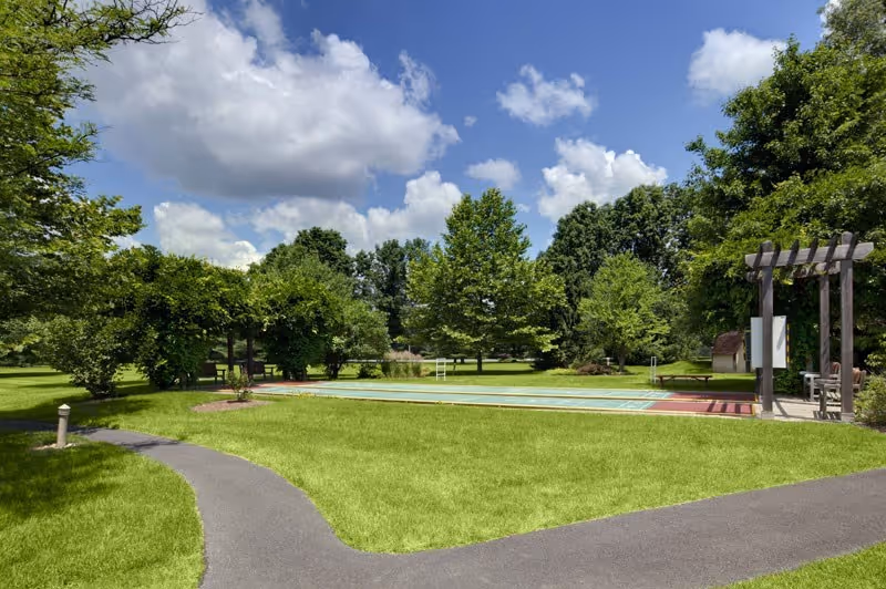 Outdoor area at Spring House Estates featuring a shuffleboard court surrounded by green grass, trees, and a paved walking path under a partly cloudy blue sky.