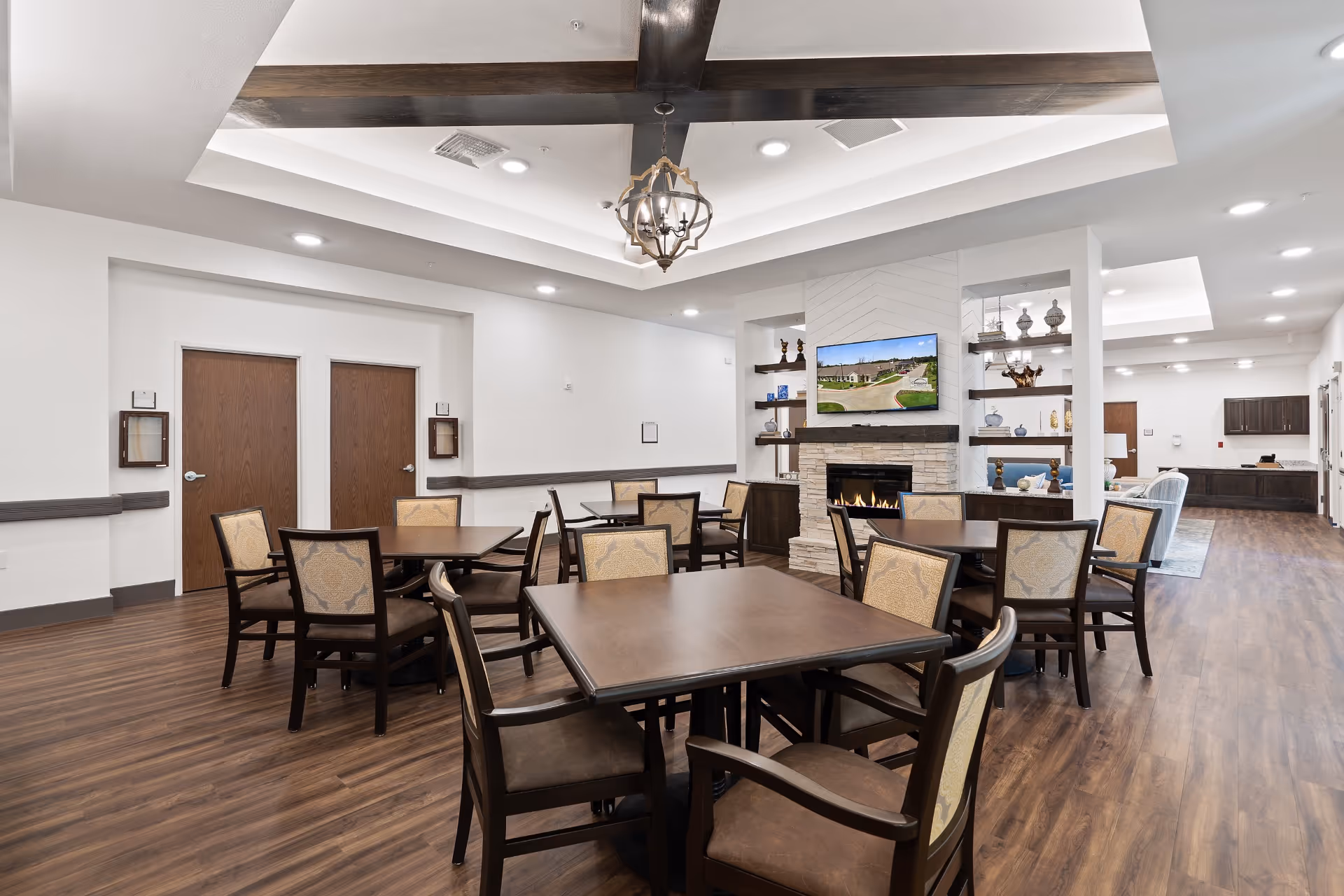 A spacious senior living common area with several wooden tables and cushioned chairs arranged on a wood floor. The room features a modern fireplace with a TV mounted above it, decorative shelves with ornaments, and a ceiling with wooden beams and a chandelier. The walls are white, and there are two closed wooden doors on one side.