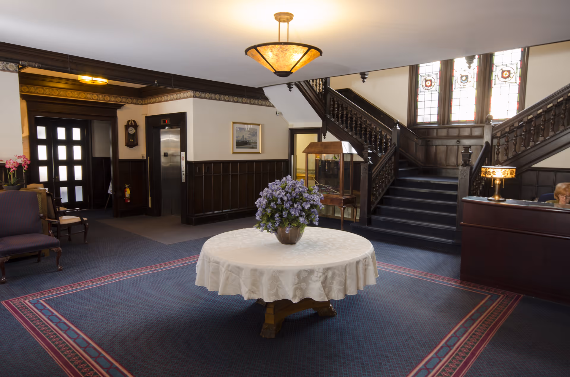Interior view of a senior living facility lobby with a round table covered with a white tablecloth and a vase of purple flowers in the center. There is a dark wooden staircase with stained glass windows behind it, an elevator, a reception desk with a lamp, and seating areas with chairs. The floor is carpeted with a patterned border.