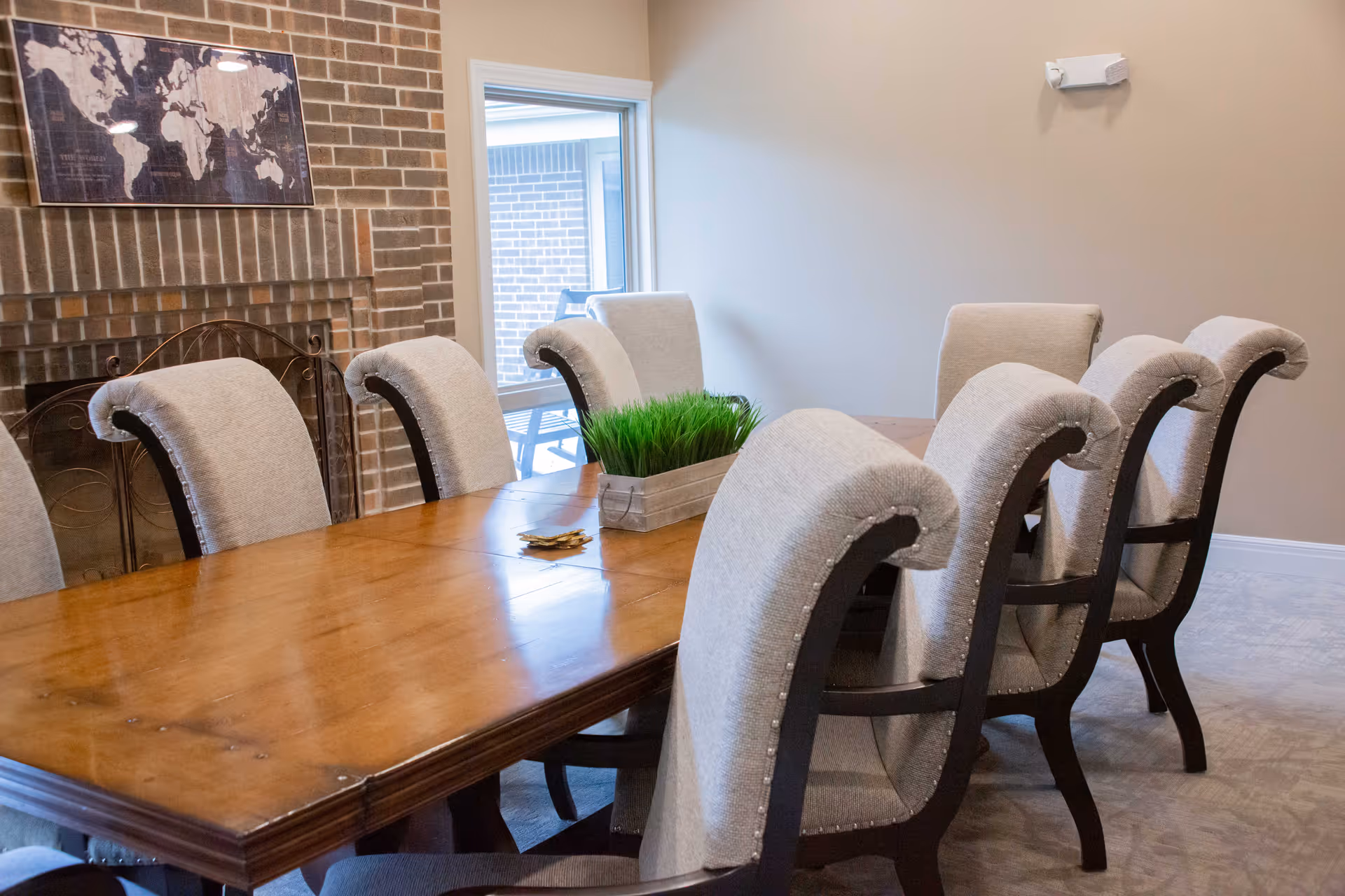 A dining room with a large wooden table surrounded by eight upholstered chairs. A rectangular planter with green grass sits in the center of the table. The room has beige walls, a brick fireplace with a metal screen, and a framed world map above the fireplace. There is a window showing an outdoor patio area with chairs.