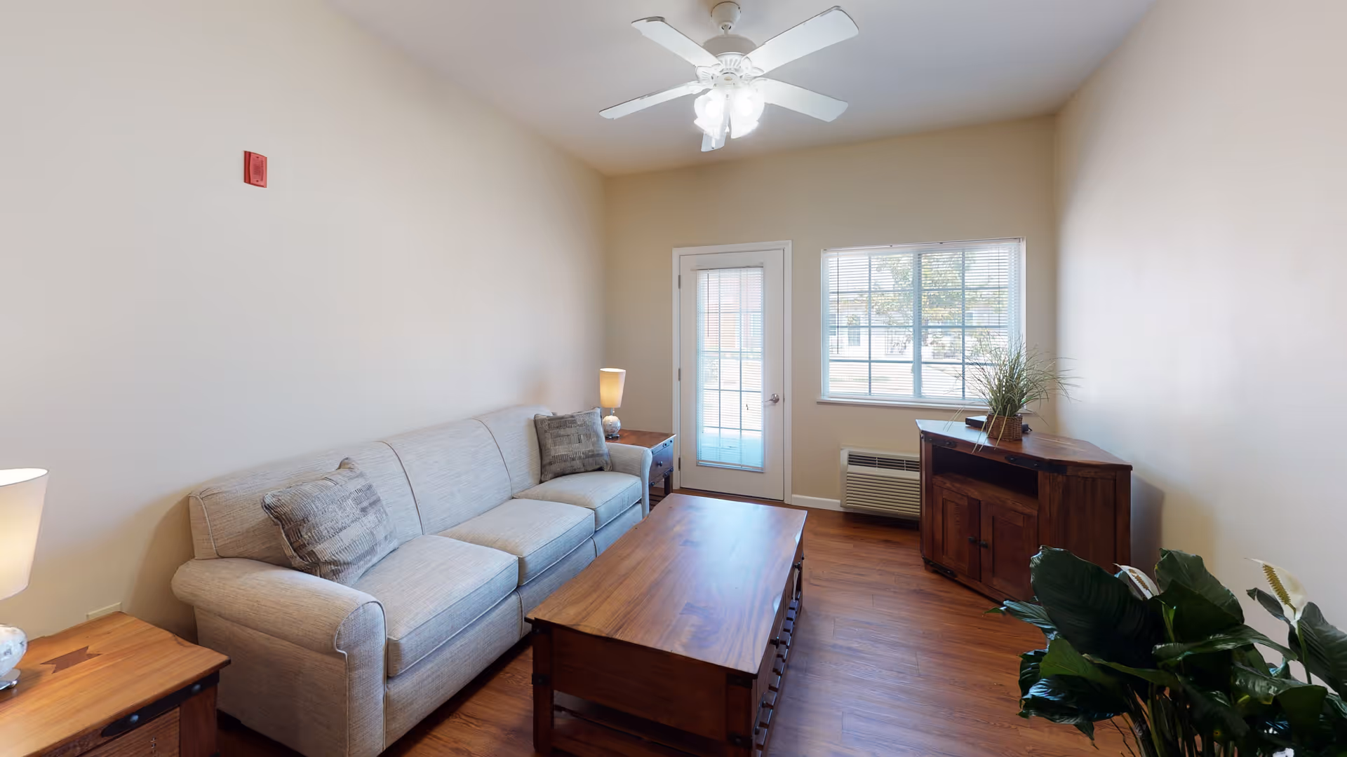 A cozy living room with a beige sofa, two side tables with lamps, a wooden coffee table, a wooden corner cabinet with a plant on top, a ceiling fan with lights, a window, and a glass door leading outside.