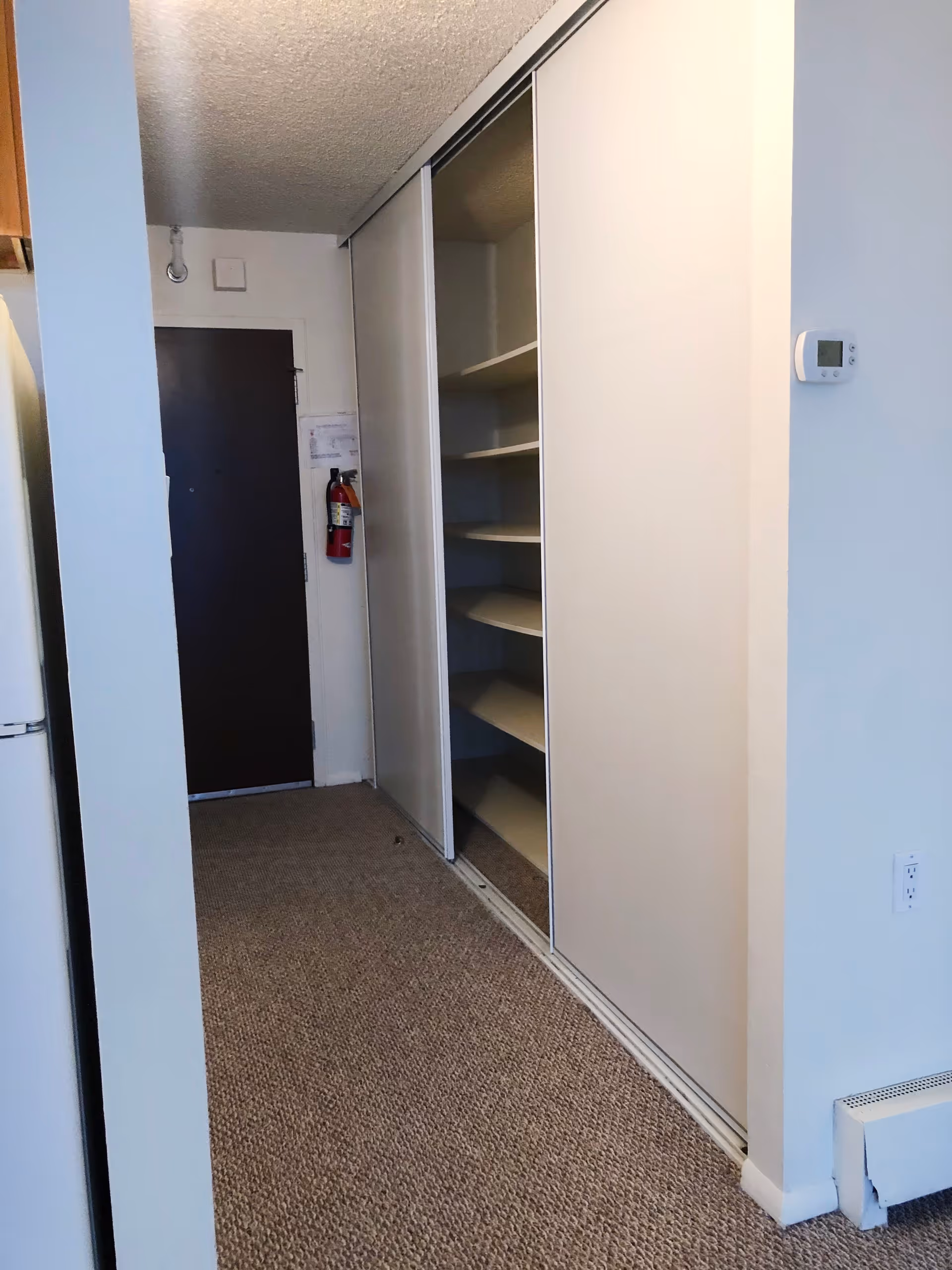 Interior hallway area with beige carpet, white walls, a dark brown door at the end, a fire extinguisher mounted on the wall, and a large closet with sliding doors partially open revealing shelves inside. A thermostat is mounted on the right wall and a white refrigerator is partially visible on the left.