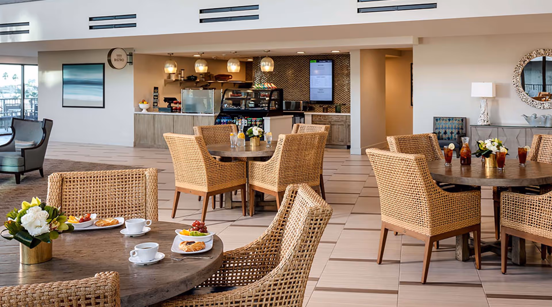 A bright and modern dining area with round wooden tables and wicker chairs. Some tables have plates with pastries, fruit, and cups of coffee, while others have glasses of iced tea and a pitcher. In the background, there is a small bistro counter with a display case, hanging pendant lights, and a digital menu screen on the wall. The space has light-colored tiled floors and neutral walls with decorative elements including a round mirror and a framed picture.