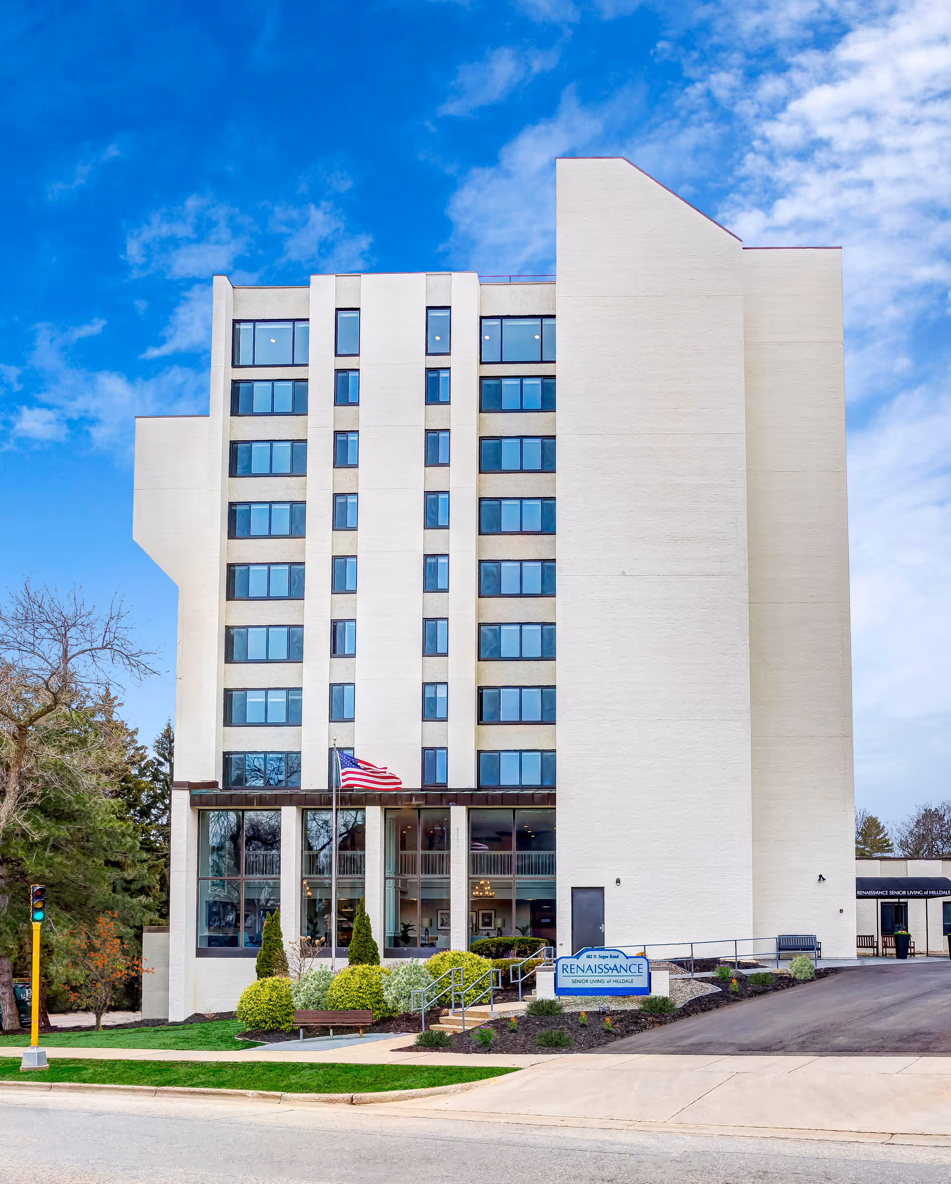 Exterior view of a multi-story senior living facility building with large windows, an American flag, landscaped bushes, and a sign that reads Renaissance Senior Living of Hillsdale. The sky is blue with some clouds.