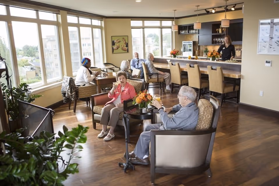 A bright and spacious common area in a senior living facility with large windows letting in natural light. Several elderly residents are seated comfortably in armchairs and at tables, enjoying drinks and conversation. A staff member stands behind a bar counter with stools, smiling and engaging with the residents. The room features wooden flooring, plants, and warm decor including flower arrangements and artwork on the walls.