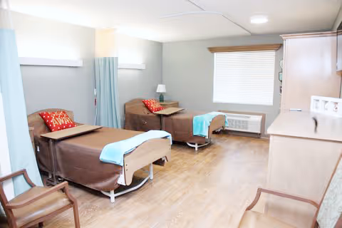 A bright shared patient room with two hospital-style beds, wood flooring, a window, and bedside furniture.